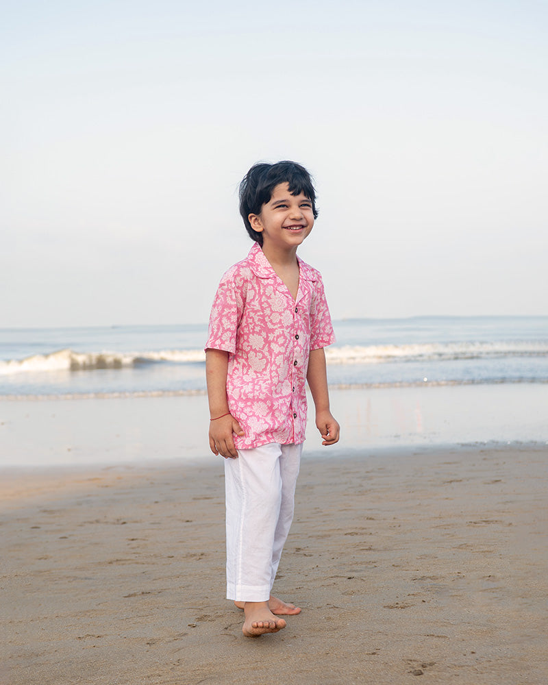 Little boy on the beach in the comfiest resort wear. Wearing a light pink floral loose fit relaxed mul mul shirt for boys with a stylish cuban collar and natural coconut shell buttons made by handblock print artisans in Jaipur, Rajasthan.