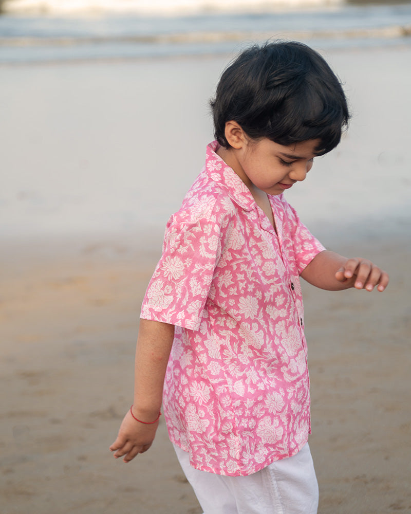 Little boy on the beach in the comfiest resort wear. Wearing a light pink floral loose fit relaxed mul mul shirt for boys with a stylish cuban collar and natural coconut shell buttons made by handblock print artisans in Jaipur, Rajasthan.