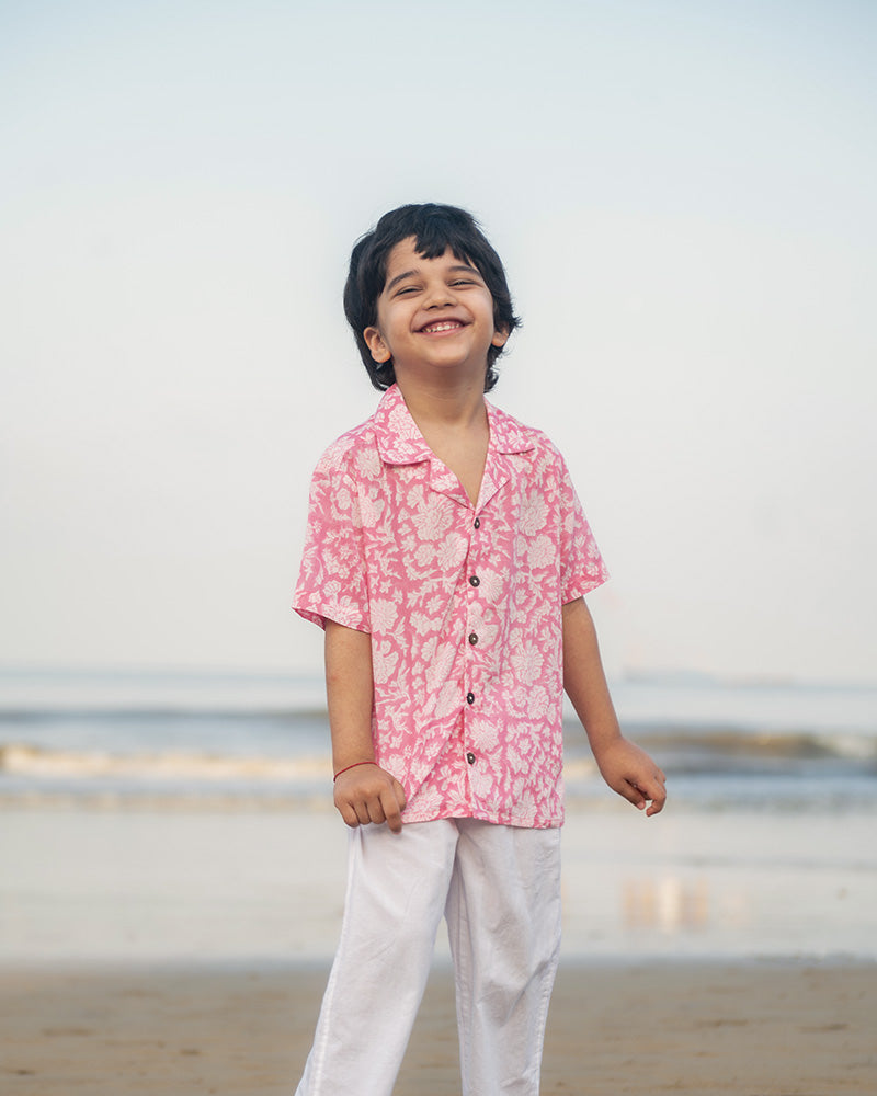Little boy on the beach in the comfiest resort wear. Wearing a light pink floral loose fit relaxed mul mul shirt for boys with a stylish cuban collar and natural coconut shell buttons made by handblock print artisans in Jaipur, Rajasthan.