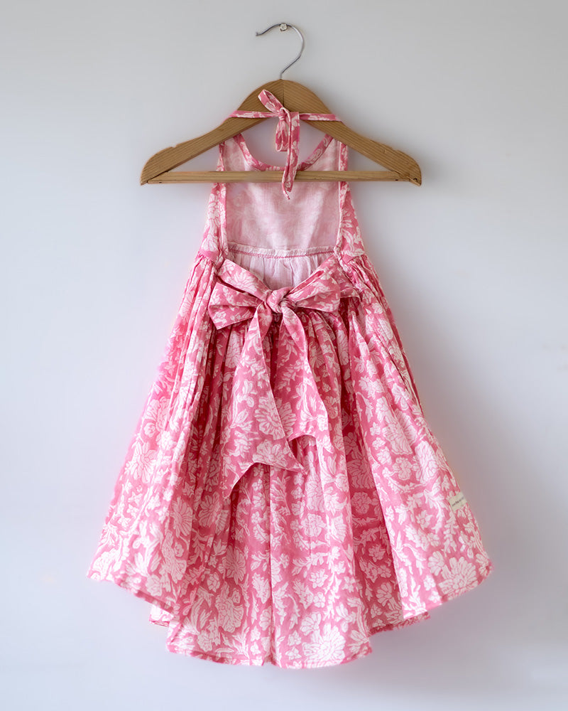 Girl on the beach in a summery pink dress. Light pink hand block printed floral summer halter neck dress made with 100% soft and breathable mulmul cotton with adjustable tie ups and elastication. Lined with mul. Upcycled hair clips in kora cotton on the little girls hair.