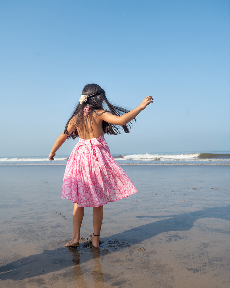 Girl on the beach in a summery pink dress. Light pink hand block printed floral summer halter neck dress made with 100% soft and breathable mulmul cotton with adjustable tie ups and elastication. Lined with mul. Upcycled hair clips in kora cotton on the little girls hair.
