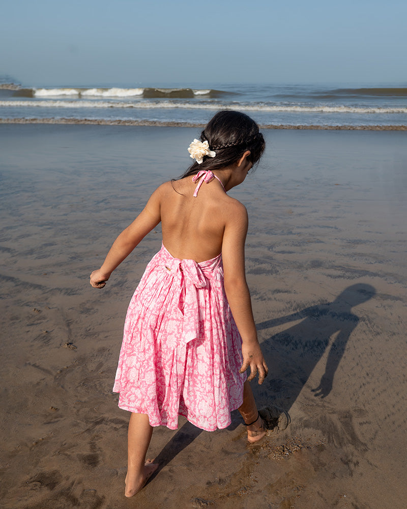 Girl on the beach in a summery pink dress. Light pink hand block printed floral summer halter neck dress made with 100% soft and breathable mulmul cotton with adjustable tie ups and elastication. Lined with mul. Upcycled hair clips in kora cotton on the little girls hair.