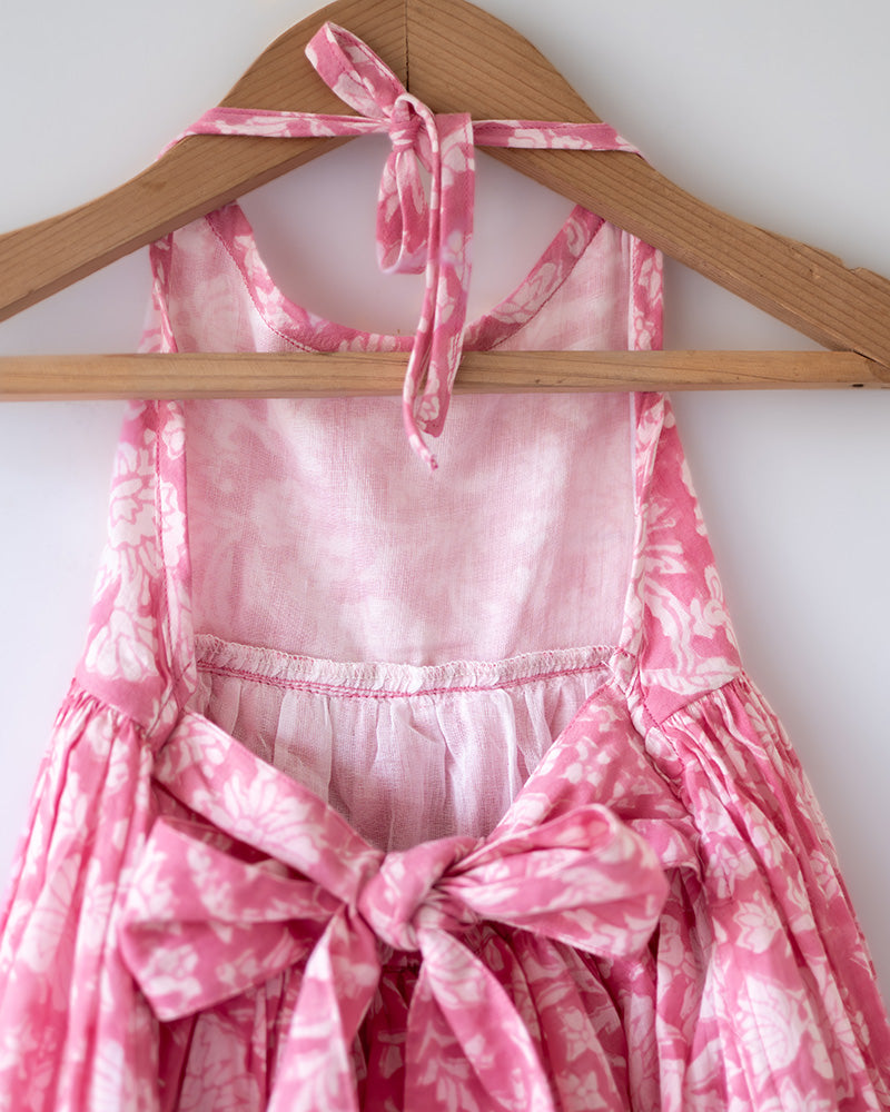 Girl on the beach in a summery pink dress. Light pink hand block printed floral summer halter neck dress made with 100% soft and breathable mulmul cotton with adjustable tie ups and elastication. Lined with mul. Upcycled hair clips in kora cotton on the little girls hair.