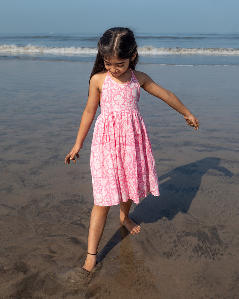 Girl on the beach in a summery pink dress. Light pink hand block printed floral summer halter neck dress made with 100% soft and breathable mulmul cotton with adjustable tie ups and elastication. Lined with mul. Upcycled hair clips in kora cotton on the little girls hair.