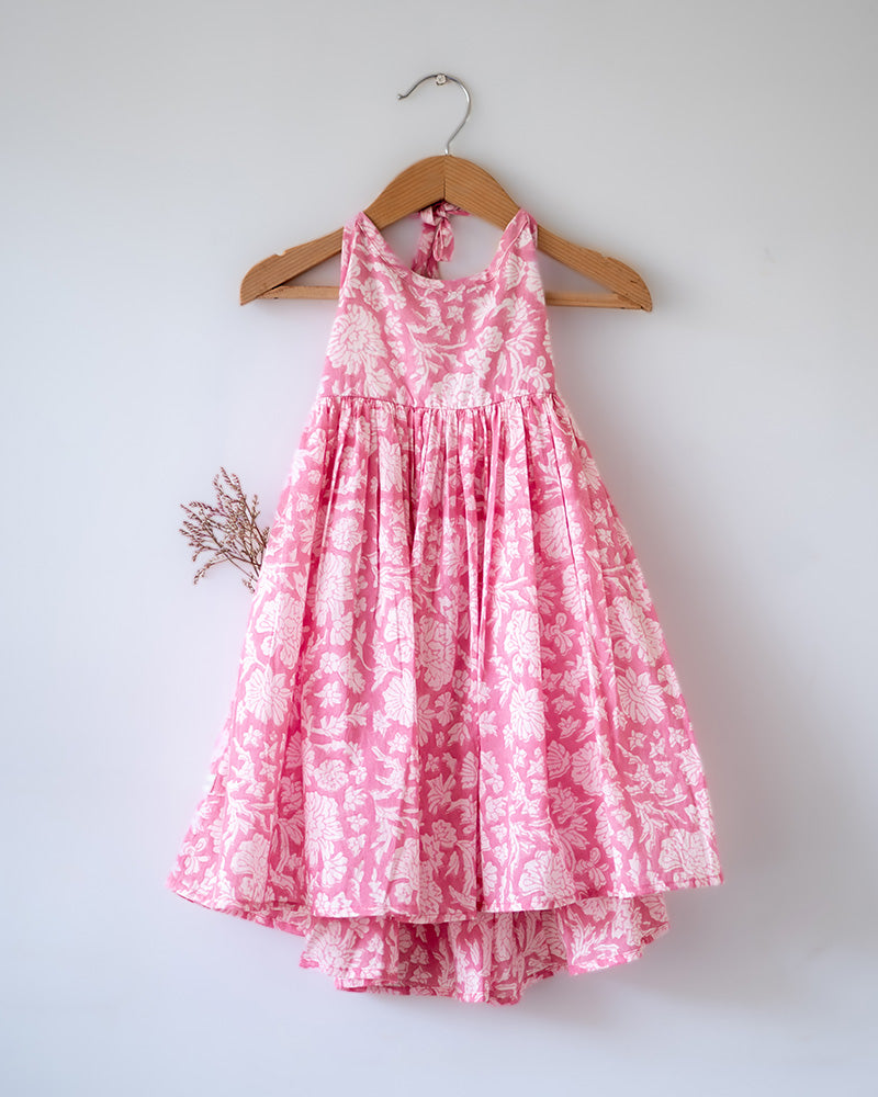 Girl on the beach in a summery pink dress. Light pink hand block printed floral summer halter neck dress made with 100% soft and breathable mulmul cotton with adjustable tie ups and elastication. Lined with mul. Upcycled hair clips in kora cotton on the little girls hair.