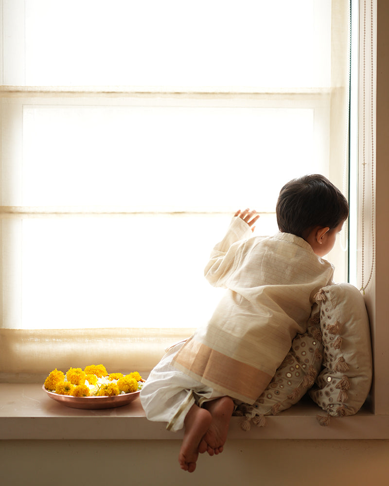 A Boy wearing an off white Indian festive Kurta with golden stripes made with Handwoven cotton silk fabric woven by artisans in Maheshwar Madhya Pradesh. This kids ethnic kurta is best for upcoming festivities like kids Navratri outfit, kids durga pooja kurta, Dussehra, dushehra, ethnic dress for Diwali, bhai dooj and kids/ boys kurta for weddings. Paired with an off white dhoti/ mundu. Perfect Kids silk wear, ethnic wear for kids, festive clothing for kids.