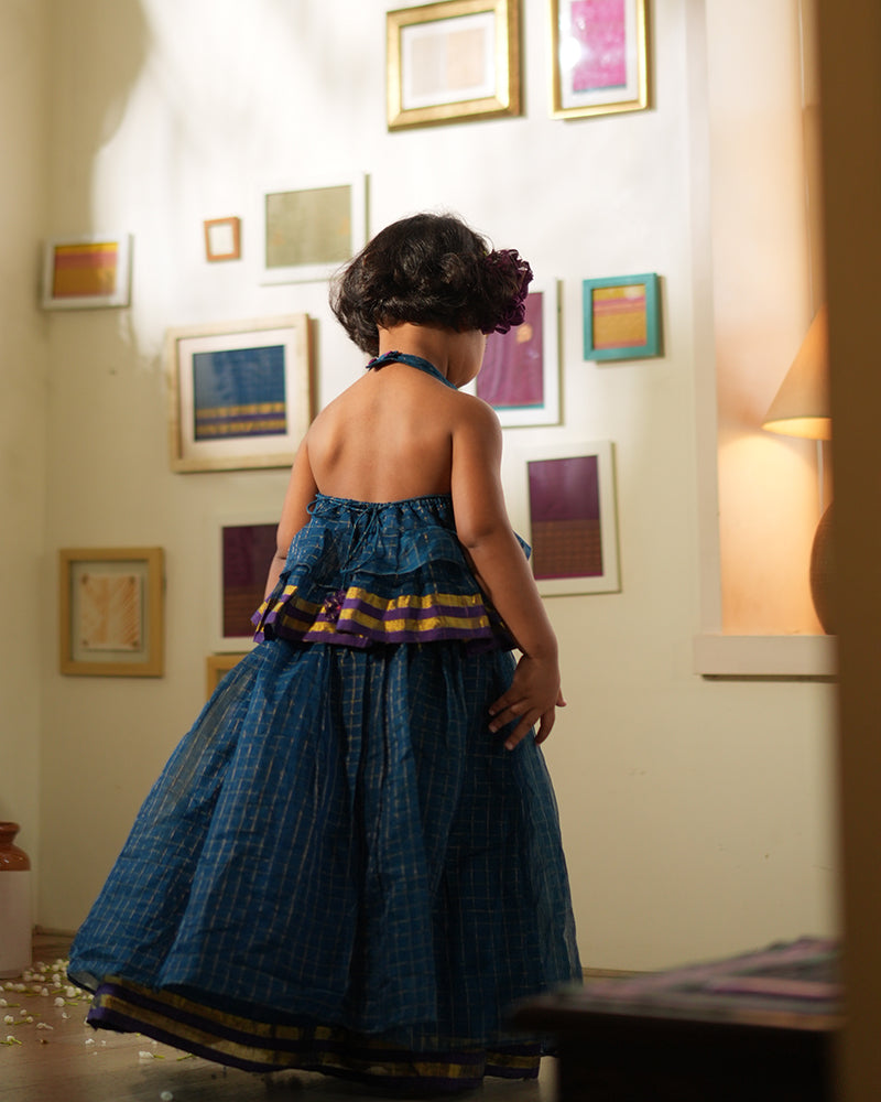 A girl wearing a deep teal blue colored cotton silk lehenga halter choli with golden checks handwoven by weavers in Maheshwar, Madhya Pradesh. This Maheshwari Cotton silk lehenga set is perfect for festivities like Navratri, durga pooja, Dussehra, dushehra, diwali pooja, bhai dooj and weddings. Gathered skirt with a ghera blouse. Kids ethnic outfits, traditional clothing for kids. Silkwear for kids. Ghera skirt