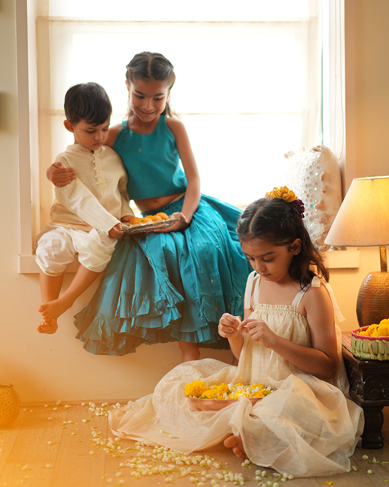 A girl wearing a bright turquoise blue colored cotton silk lehenga choli with golden buttis handwoven by weavers in Maheshwar, Madhya Pradesh. This Maheshwari Cotton silk lehenga set is perfect for festivities like Navratri, durga pooja, Dussehra, dushehra, diwali pooja, bhai dooj and weddings. Celebrating diwali with siblings. Kids ethnic wear, kids traditional clothing. Kids festive wear. Kids silk wear.