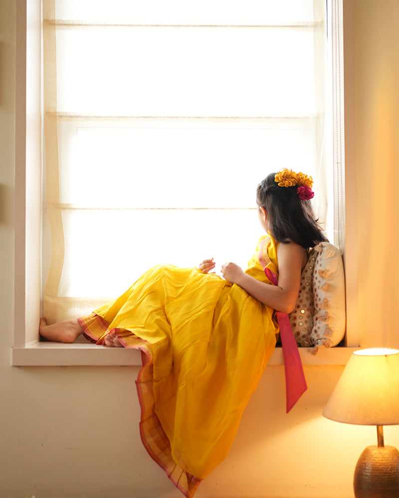 A girl wearing a bright yellow colored cotton silk lehenga handwoven by weavers in Maheshwar, Madhya Pradesh. This Maheshwari Cotton silk lehenga set is perfect for festivities like Navratri, durga pooja, Dussehra, dushehra, diwali pooja, bhai dooj and weddings. Kids ethnic outfits, traditional clothing for kids. Silkwear for kids.