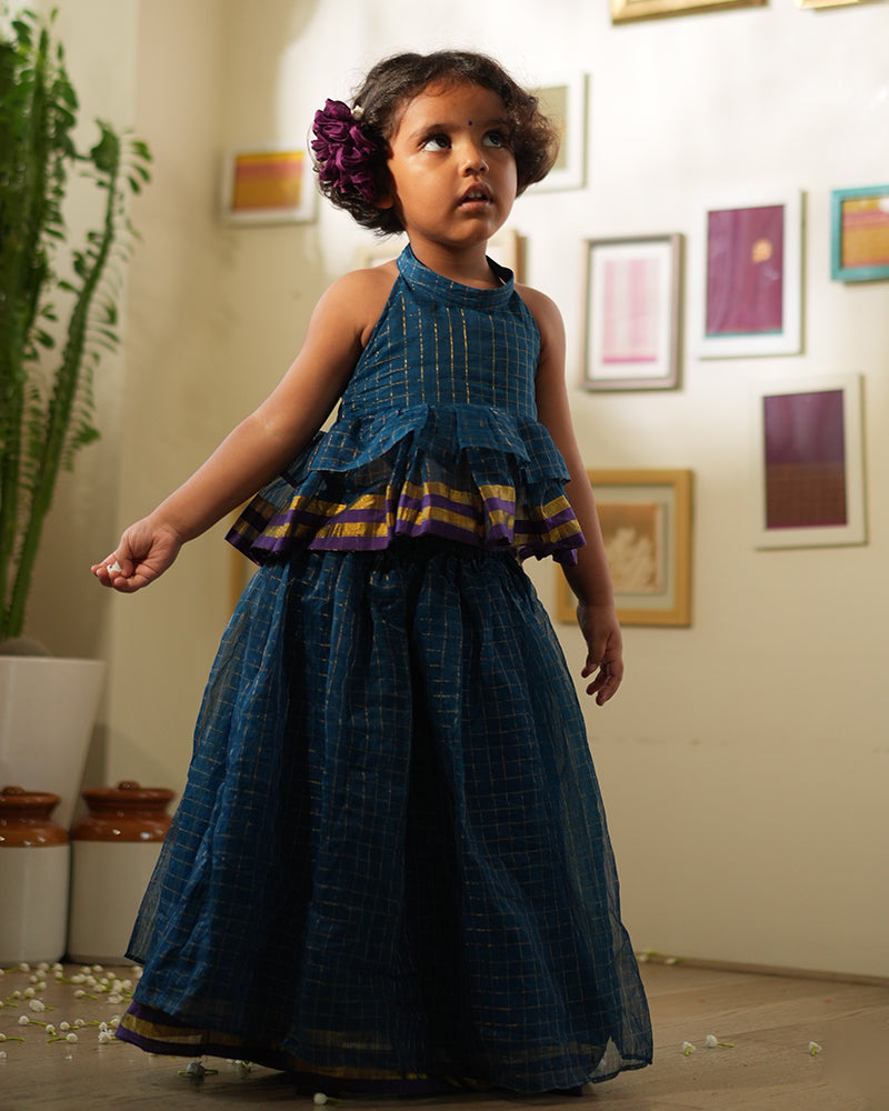 A girl wearing a deep teal blue colored cotton silk lehenga halter choli with golden checks handwoven by weavers in Maheshwar, Madhya Pradesh. This Maheshwari Cotton silk lehenga set is perfect for festivities like Navratri, durga pooja, Dussehra, dushehra, diwali pooja, bhai dooj and weddings. Gathered skirt with a ghera blouse. Kids ethnic outfits, traditional clothing for kids. Silkwear for kids. Ghera skirt