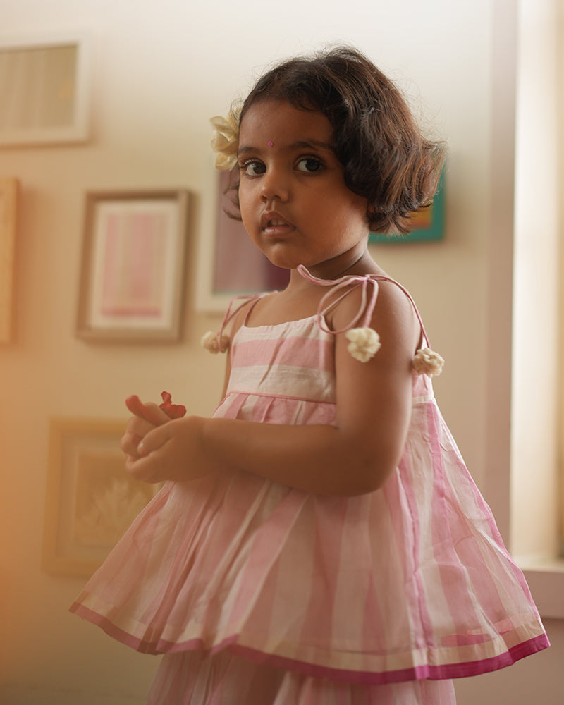A girl wearing a cute baby pink and white striped cotton silk lehenga handwoven by weavers in Maheshwar, Madhya Pradesh. This Maheshwari Cotton silk lehenga set is perfect for festivities like Navratri, durga pooja, Dussehra, dushehra, diwali pooja, bhai dooj and weddings. Kids ethnic outfits, traditional clothing for kids. Silkwear for kids.
