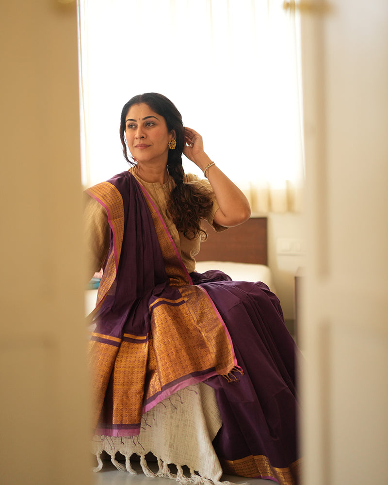 A woman wearing a deep purple wine colored cotton silk saree handwoven by weavers in Maheshwar, Madhya Pradesh. This Maheshwari Cotton silk saree is perfect for festivities like Navratri, durga pooja, Dussehra, dushehra, diwali pooja, bhai dooj and weddings. Paired with a golden blouse. Women's silk wear, silk saree for women, cotton silk saree, heavy pallu border sarees.