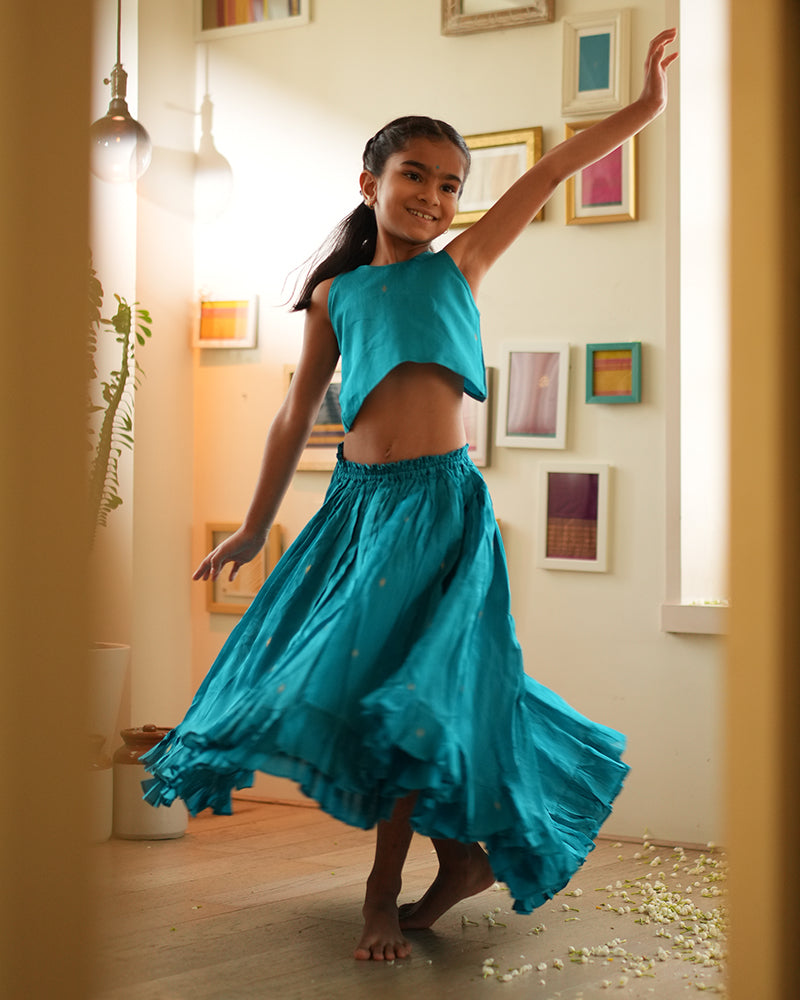 A girl wearing a bright turquoise blue colored cotton silk lehenga choli with golden buttis handwoven by weavers in Maheshwar, Madhya Pradesh. This Maheshwari Cotton silk lehenga set is perfect for festivities like Navratri, durga pooja, Dussehra, dushehra, diwali pooja, bhai dooj and weddings. High low skirt with a crop top blouse. Kids ethnic outfits, traditional clothing for kids. Silkwear for kids.