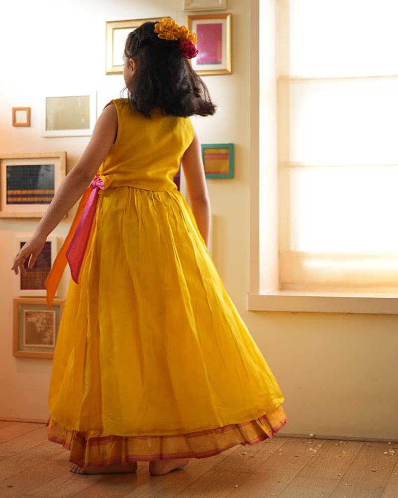 A girl wearing a bright yellow colored cotton silk lehenga handwoven by weavers in Maheshwar, Madhya Pradesh. This Maheshwari Cotton silk lehenga set is perfect for festivities like Navratri, durga pooja, Dussehra, dushehra, diwali pooja, bhai dooj and weddings. Kids ethnic outfits, traditional clothing for kids. Silkwear for kids.