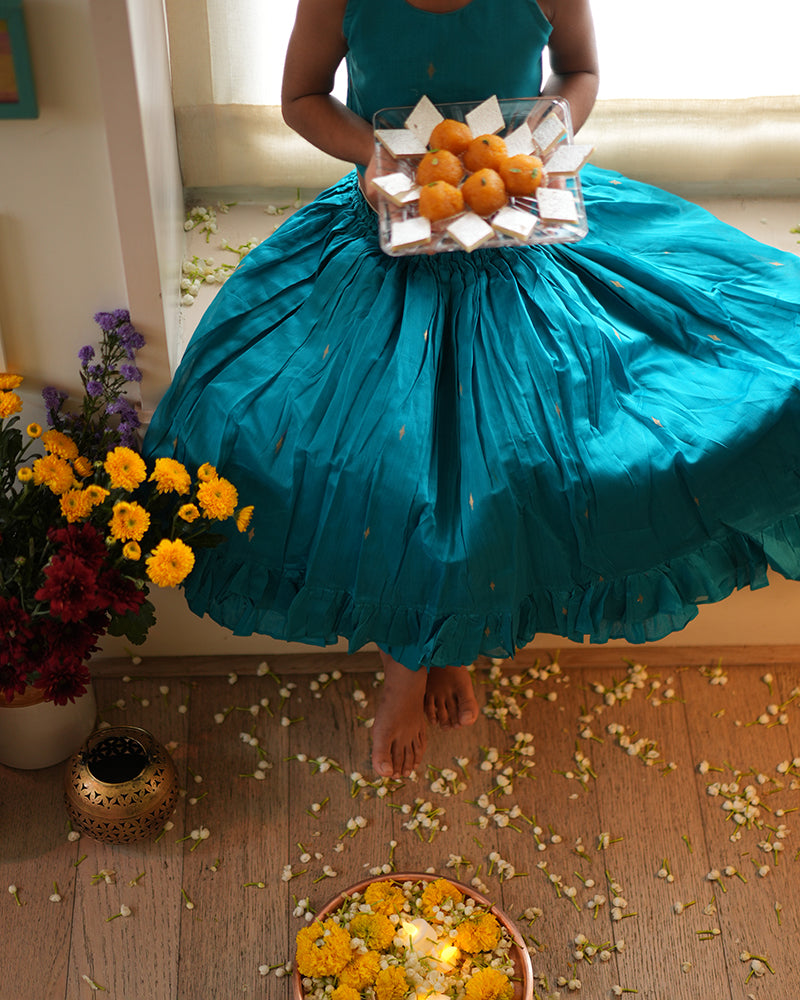 A girl wearing a bright turquoise blue colored cotton silk lehenga choli with golden buttis handwoven by weavers in Maheshwar, Madhya Pradesh. This Maheshwari Cotton silk lehenga set is perfect for festivities like Navratri, durga pooja, Dussehra, dushehra, diwali pooja, bhai dooj and weddings. Celebrating diwali.