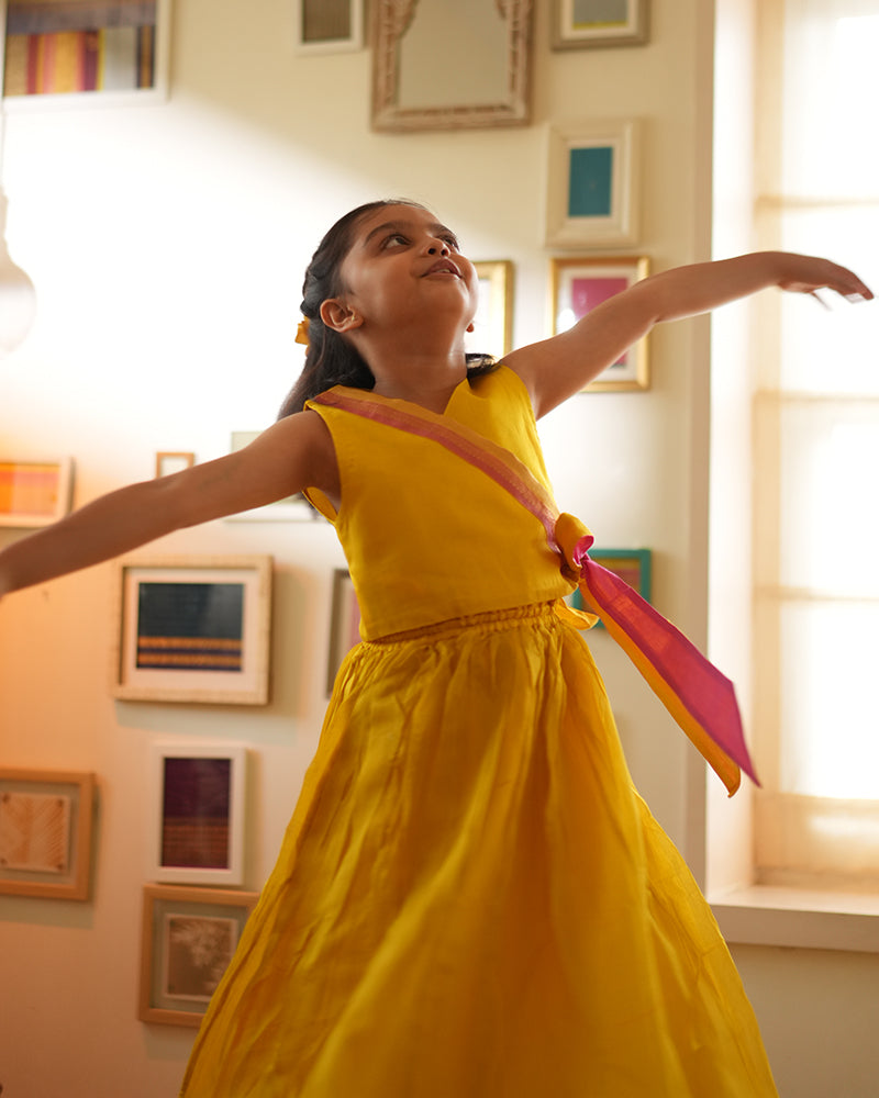 A girl wearing a bright yellow colored cotton silk lehenga handwoven by weavers in Maheshwar, Madhya Pradesh. This Maheshwari Cotton silk lehenga set is perfect for festivities like Navratri, durga pooja, Dussehra, dushehra, diwali pooja, bhai dooj and weddings. Kids ethnic outfits, traditional clothing for kids. Silkwear for kids.