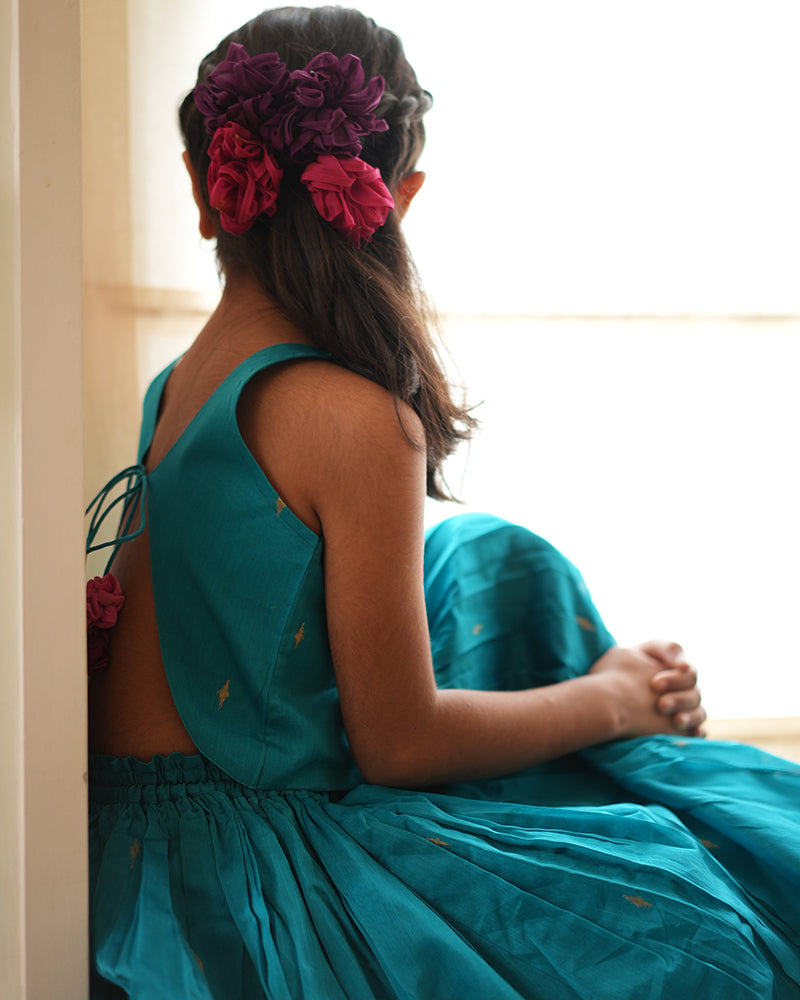 A girl wearing a bright turquoise blue colored cotton silk lehenga choli with golden buttis handwoven by weavers in Maheshwar, Madhya Pradesh. This Maheshwari Cotton silk lehenga set is perfect for festivities like Navratri, durga pooja, Dussehra, dushehra, diwali pooja, bhai dooj and weddings. Tie back dori blouse. Kids ethnic wear, kids traditional outfits. Girls ghagra. Floral hair accessories.