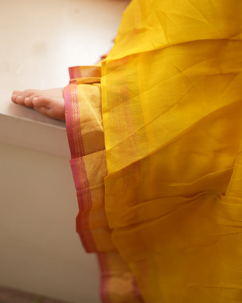 A girl wearing a bright yellow colored cotton silk lehenga handwoven by weavers in Maheshwar, Madhya Pradesh. Ghagra has pink border and crop blouse has cross pattern tie up. This Maheshwari Cotton silk lehenga set is perfect for festivities like Navratri, durga pooja, Dussehra, dushehra, diwali pooja, bhai dooj and weddings. Kids ethnic outfits, traditional clothing for kids. Silkwear for kids.