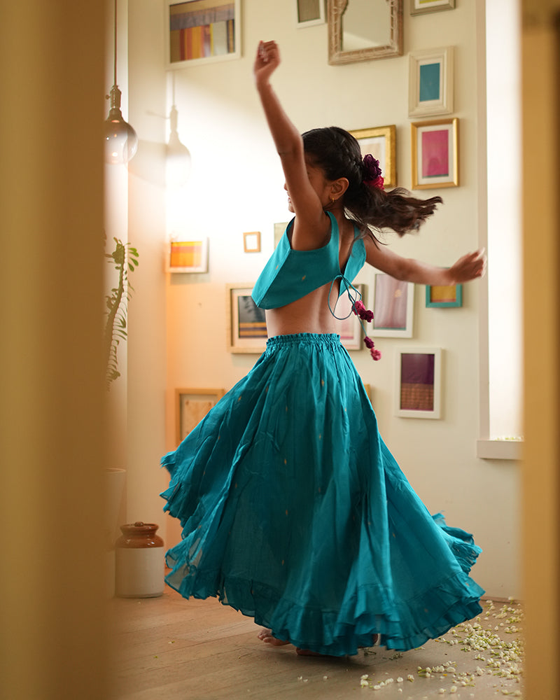 A girl wearing a bright turquoise blue colored cotton silk lehenga choli with golden buttis handwoven by weavers in Maheshwar, Madhya Pradesh. This Maheshwari Cotton silk lehenga set is perfect for festivities like Navratri, durga pooja, Dussehra, dushehra, diwali pooja, bhai dooj and weddings. High low skirt with a crop top blouse. Kids silk wear. Kids ethnic wear.