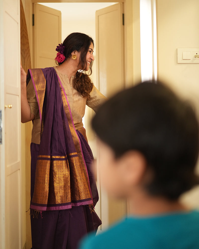 A woman wearing a deep purple wine colored cotton silk saree handwoven by weavers in Maheshwar, Madhya Pradesh. This Maheshwari Cotton silk saree is perfect for festivities like Navratri, durga pooja, Dussehra, dushehra, diwali pooja, bhai dooj and weddings. Paired with a golden blouse. Women's silk wear, silk saree for women, cotton silk saree, heavy pallu border sarees.