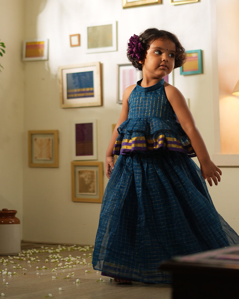 A girl wearing a deep teal blue colored cotton silk lehenga halter choli with golden checks handwoven by weavers in Maheshwar, Madhya Pradesh. This Maheshwari Cotton silk lehenga set is perfect for festivities like Navratri, durga pooja, Dussehra, dushehra, diwali pooja, bhai dooj and weddings. Gathered skirt with a ghera blouse. Kids ethnic outfits, traditional clothing for kids. Silkwear for kids. Ghera skirt
