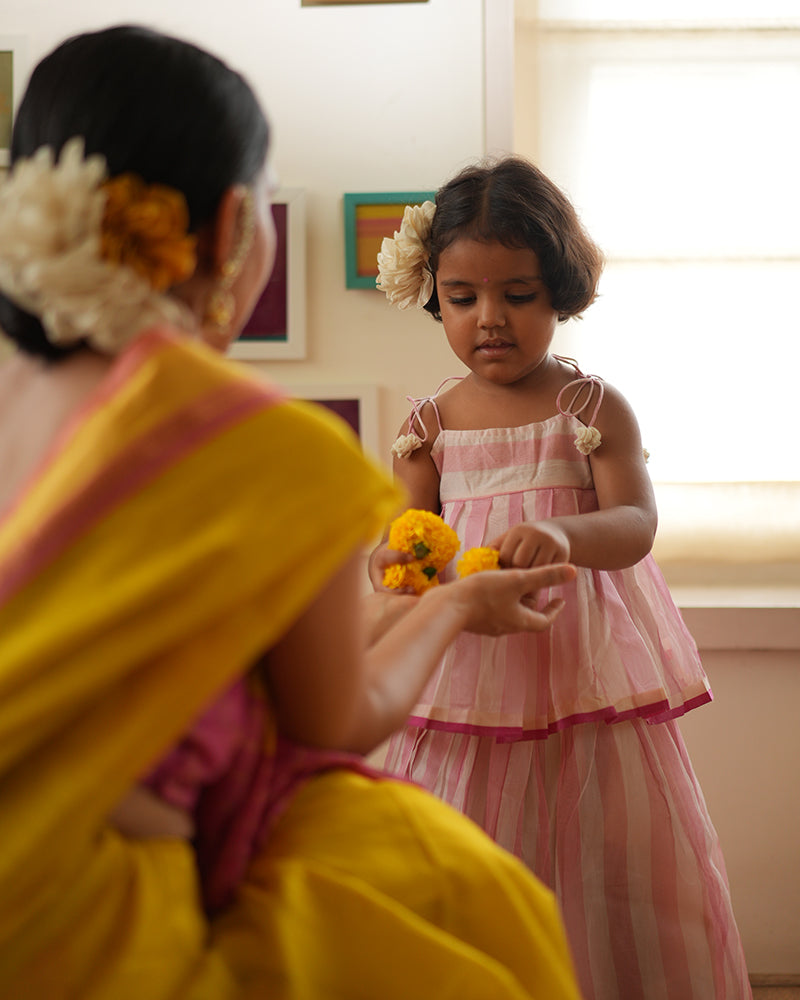 A girl wearing a cute baby pink and white striped cotton silk lehenga handwoven by weavers in Maheshwar, Madhya Pradesh. This Maheshwari Cotton silk lehenga set is perfect for festivities like Navratri, durga pooja, Dussehra, dushehra, diwali pooja, bhai dooj and weddings. Kids ethnic outfits, traditional clothing for kids. Silkwear for kids.