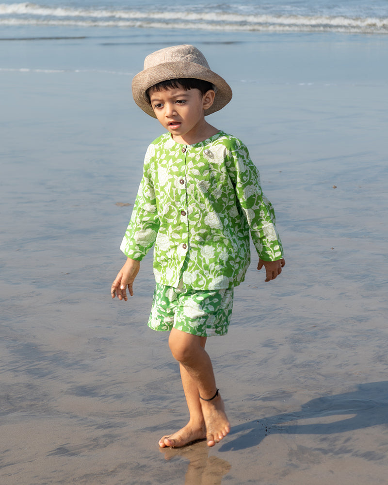 A cute boy with a sun hat on the beach wearing the unisex vintage print forest green colour floral kimono shirt and shorts coord set for girls and boys in breathable mulmul cotton that is hand block printed in Jaipur. Shorts have a soft elastic waist, coconut shell buttons and is handmade in India