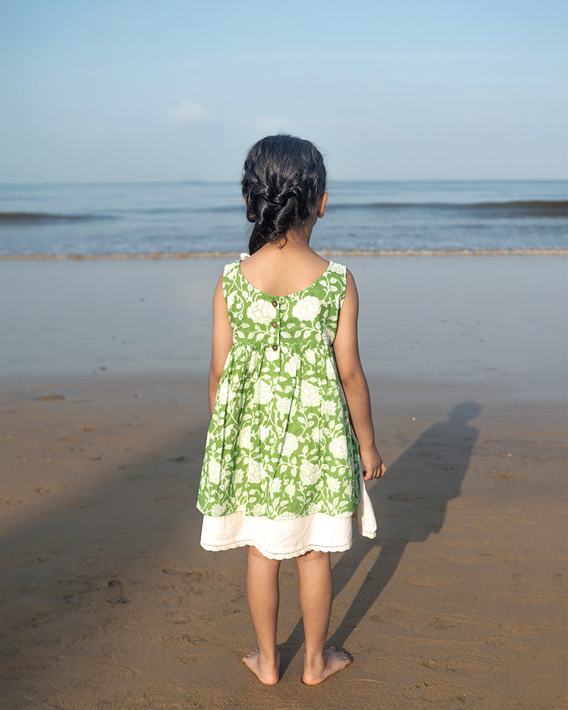 Little girl twirling in a sleeveless mul mul cotton layered pinafore style cute summer frock in forest green and white colour on the beach. Vintage crochet detailing at the bottom of the dress. 