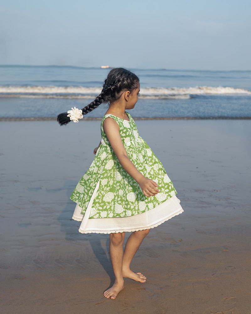 Little girl twirling in a sleeveless mul mul cotton layered pinafore style cute summer frock in forest green and white colour on the beach. Vintage crochet detailing at the bottom of the dress. 