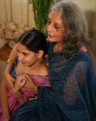 A woman wearing a deep teal blue colored cotton silk saree with golden checks handwoven by weavers in Maheshwar, Madhya Pradesh. This Maheshwari Cotton silk saree is perfect for festivities like Navratri, durga pooja, Dussehra, dushehra, diwali pooja, bhai dooj and weddings. Women's silk wear, silk saree for women, cotton silk saree, heavy pallu border sarees. Mom & daughter duo.