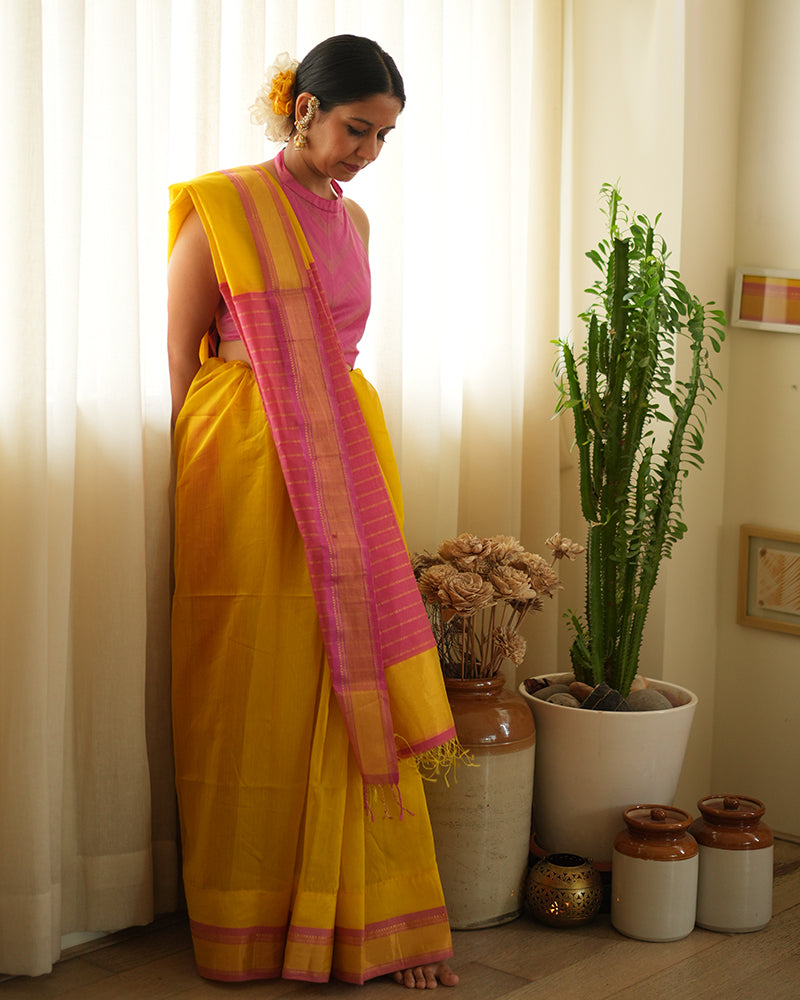 A woman wearing a bright yellow colored cotton silk saree with a pink and gold border and pallu with pink and gold stripes handwoven by weavers in Maheshwar, Madhya Pradesh. This Maheshwari Cotton silk saree is perfect for festivities like Navratri, durga pooja, Dussehra, dushehra, diwali pooja, bhai dooj and weddings. Women's silk wear, silk saree for women, cotton silk saree, heavy pallu sarees. 