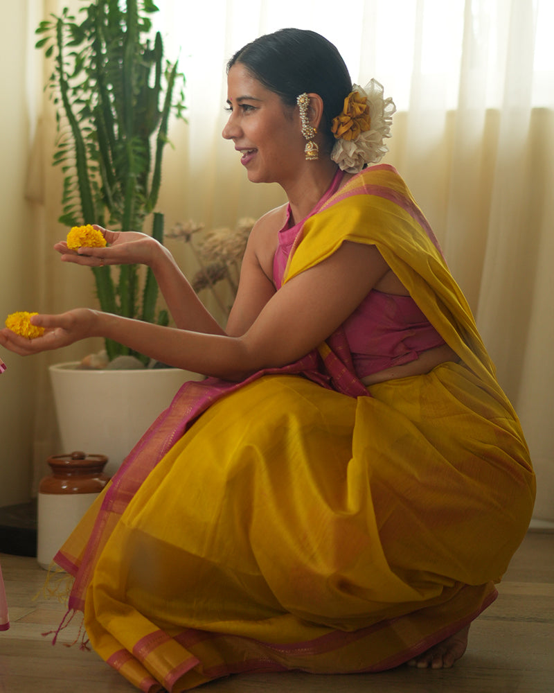 A woman wearing a bright yellow colored cotton silk saree with a pink and gold border and pallu with pink and gold stripes handwoven by weavers in Maheshwar, Madhya Pradesh. This Maheshwari Cotton silk saree is perfect for festivities like Navratri, durga pooja, Dussehra, dushehra, diwali pooja, bhai dooj and weddings. Women's silk wear, silk saree for women, cotton silk saree, heavy pallu sarees. South Indian saree, south Indian look.