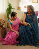 A woman wearing a bright pink colored cotton silk saree with a blue border and golden stripes handwoven by weavers in Maheshwar, Madhya Pradesh. This Maheshwari Cotton silk saree is perfect for festivities like Navratri, durga pooja, Dussehra, dushehra, diwali pooja, bhai dooj and weddings. Women's silk wear, silk saree for women, cotton silk saree. Mom and daughter duo. Mother with Daughter celebrating diwali. 