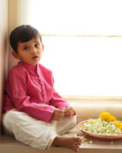 A Boy wearing a bright rani Pink Indian festive Kurta with thick golden stripes made with Handwoven cotton silk fabric woven by artisans in Maheshwar Madhya Pradesh. This kids ethnic kurta is best for upcoming festivities like kids Navratri outfit, kids durga pooja kurta, Dussehra, dushehra, ethnic dress for Diwali, bhai dooj and kids/ boys kurta for weddings. Paired with an off white dhoti/ mundu. Perfect Kids silk wear, ethnic wear for kids, festive clothing for kids.