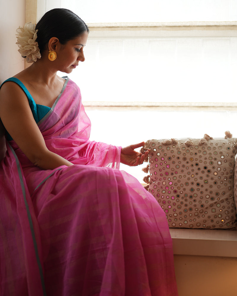 A woman wearing a bright pink colored cotton silk saree with a blue border and golden stripes handwoven by weavers in Maheshwar, Madhya Pradesh. This Maheshwari Cotton silk saree is perfect for festivities like Navratri, durga pooja, Dussehra, dushehra, diwali pooja, bhai dooj and weddings. Women's silk wear, silk saree for women, cotton silk saree. 