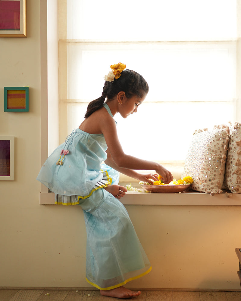A girl wearing a cute light baby blue and yellow cotton silk sharara suit set handwoven with silver polka dots by weavers in Maheshwar, Madhya Pradesh. This Maheshwari Cotton silk suit set is perfect for festivities like Navratri, durga pooja, Dussehra, dushehra, diwali pooja, bhai dooj and weddings. Kids ethnic outfits, traditional clothing for kids. Silkwear for kids. Halter neck kurti top, salwar kamiz. 
