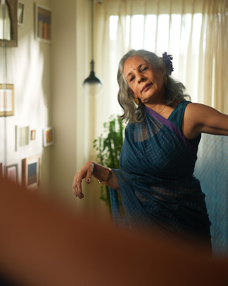 A woman wearing a deep teal blue colored cotton silk saree with golden checks handwoven by weavers in Maheshwar, Madhya Pradesh. This Maheshwari Cotton silk saree is perfect for festivities like Navratri, durga pooja, Dussehra, dushehra, diwali pooja, bhai dooj and weddings. Women's silk wear, silk saree for women, cotton silk saree, heavy pallu border sarees. 