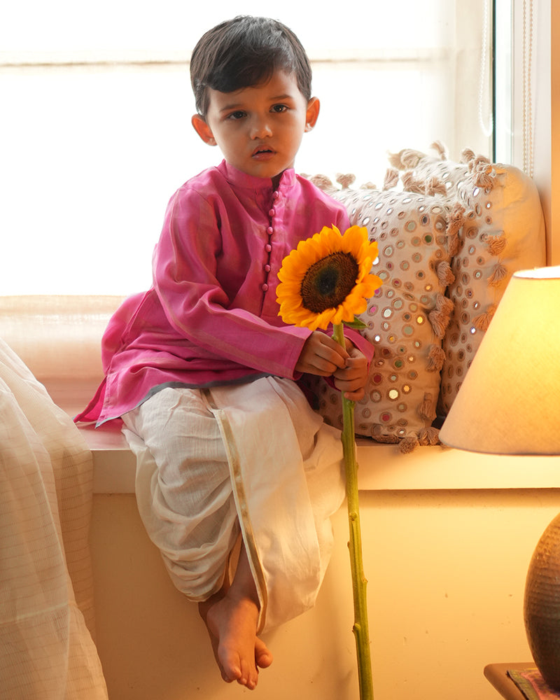 A Boy wearing a bright rani Pink Indian festive Kurta with thick golden stripes made with Handwoven cotton silk fabric woven by artisans in Maheshwar Madhya Pradesh. This kids ethnic kurta is best for upcoming festivities like kids Navratri outfit, kids durga pooja kurta, Dussehra, dushehra, ethnic dress for Diwali, bhai dooj and kids/ boys kurta for weddings. Paired with an off white dhoti/ mundu. Perfect Kids silk wear, ethnic wear for kids, festive clothing for kids.