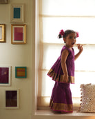 A girl wearing a cute deep purple violet cotton silk sharara suit set handwoven with golden border by weavers in Maheshwar, Madhya Pradesh. This Maheshwari Cotton silk suit set is perfect for festivities like Navratri, durga pooja, Dussehra, dushehra, diwali pooja, bhai dooj and weddings. Kids ethnic outfits, traditional clothing for kids. Silkwear for kids. Sharara set, suit set, salwar set for girls. 