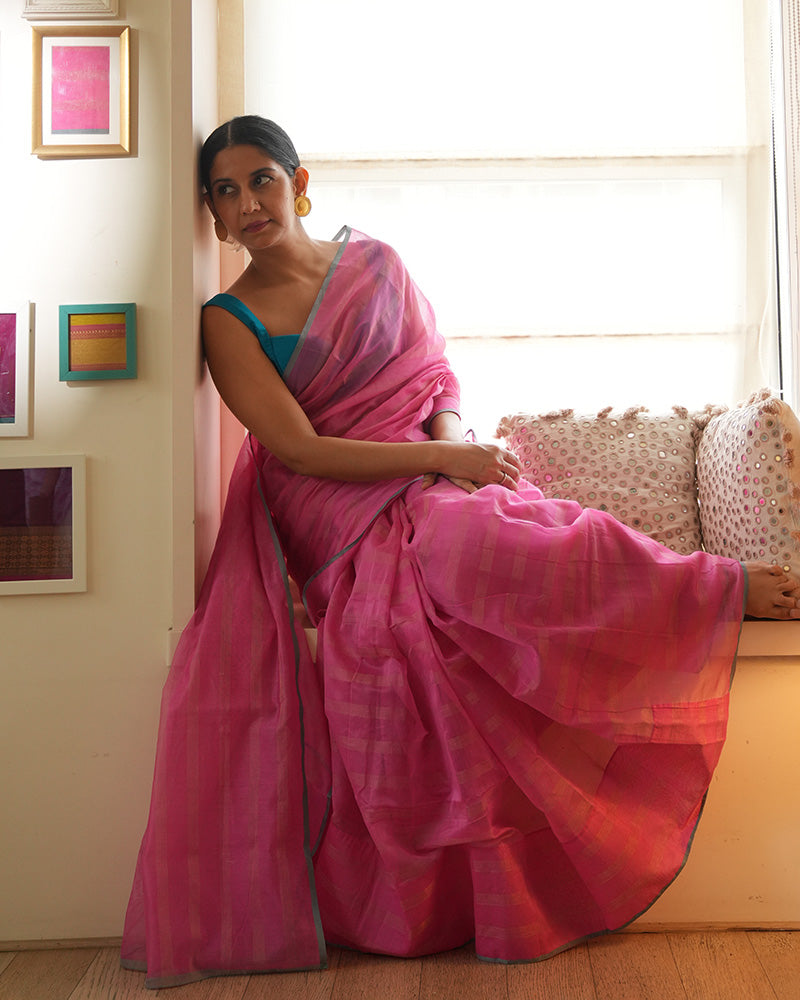 A woman wearing a bright pink colored cotton silk saree with a blue border and golden stripes handwoven by weavers in Maheshwar, Madhya Pradesh. This Maheshwari Cotton silk saree is perfect for festivities like Navratri, durga pooja, Dussehra, dushehra, diwali pooja, bhai dooj and weddings. Women's silk wear, silk saree for women, cotton silk saree. 