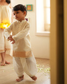 A Boy wearing an off white Indian festive Kurta with golden stripes made with Handwoven cotton silk fabric woven by artisans in Maheshwar Madhya Pradesh. This kids ethnic kurta is best for upcoming festivities like kids Navratri outfit, kids durga pooja kurta, Dussehra, dushehra, ethnic dress for Diwali, bhai dooj and kids/ boys kurta for weddings. Paired with an off white dhoti/ mundu. Perfect Kids silk wear, ethnic wear for kids, festive clothing for kids.