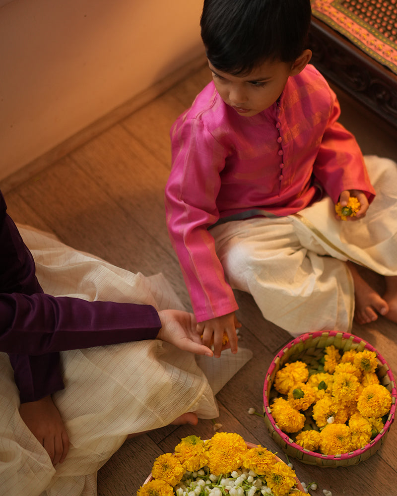 A Boy wearing a bright rani Pink Indian festive Kurta with thick golden stripes made with Handwoven cotton silk fabric woven by artisans in Maheshwar Madhya Pradesh. This kids ethnic kurta is best for upcoming festivities like kids Navratri outfit, kids durga pooja kurta, Dussehra, dushehra, ethnic dress for Diwali, bhai dooj and kids/ boys kurta for weddings. Paired with an off white dhoti/ mundu. Perfect Kids silk wear, ethnic wear for kids, festive clothing for kids. Sibling love. 