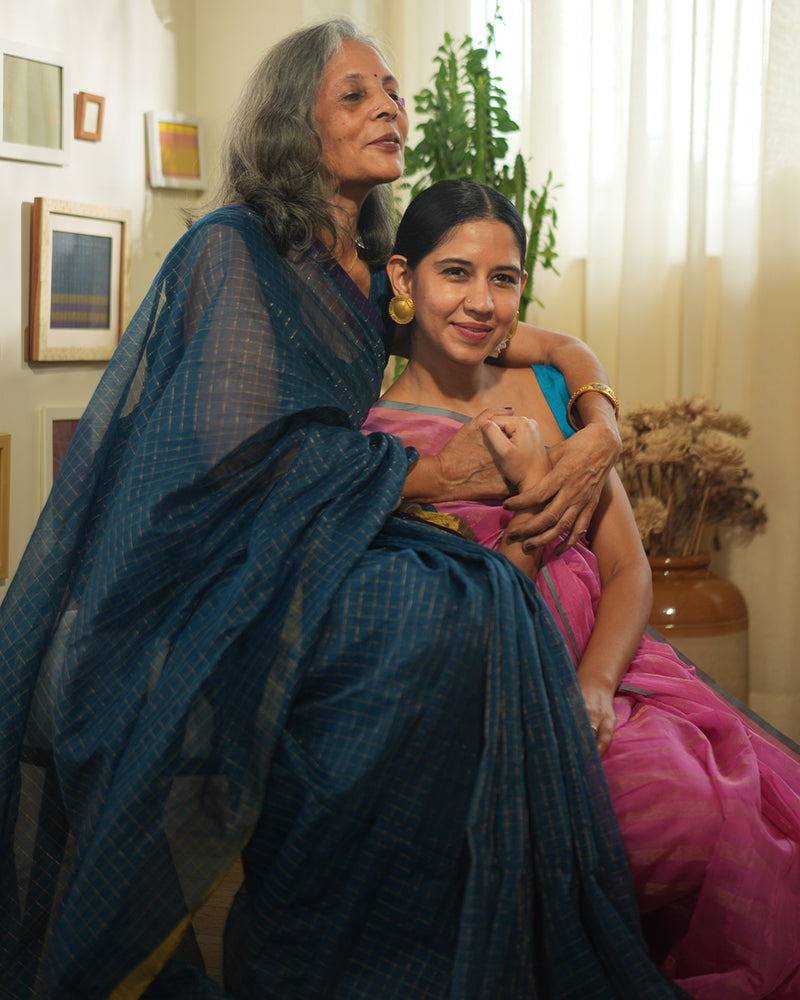 A woman wearing a deep teal blue colored cotton silk saree with golden checks handwoven by weavers in Maheshwar, Madhya Pradesh. This Maheshwari Cotton silk saree is perfect for festivities like Navratri, durga pooja, Dussehra, dushehra, diwali pooja, bhai dooj and weddings. Women's silk wear, silk saree for women, cotton silk saree, heavy pallu border sarees. 