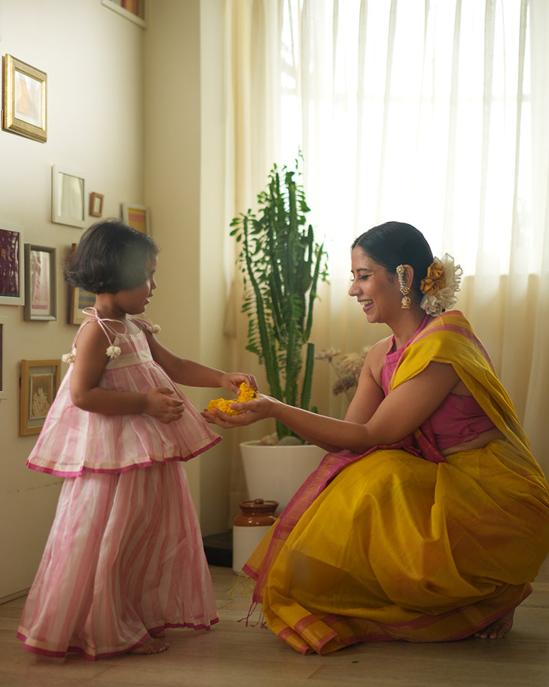 A woman wearing a bright yellow colored cotton silk saree with a pink and gold border and pallu with pink and gold stripes handwoven by weavers in Maheshwar, Madhya Pradesh. This Maheshwari Cotton silk saree is perfect for festivities like Navratri, durga pooja, Dussehra, dushehra, diwali pooja, bhai dooj and weddings. Women's silk wear, silk saree for women, cotton silk saree, heavy pallu sarees. Mom daughter duo. 