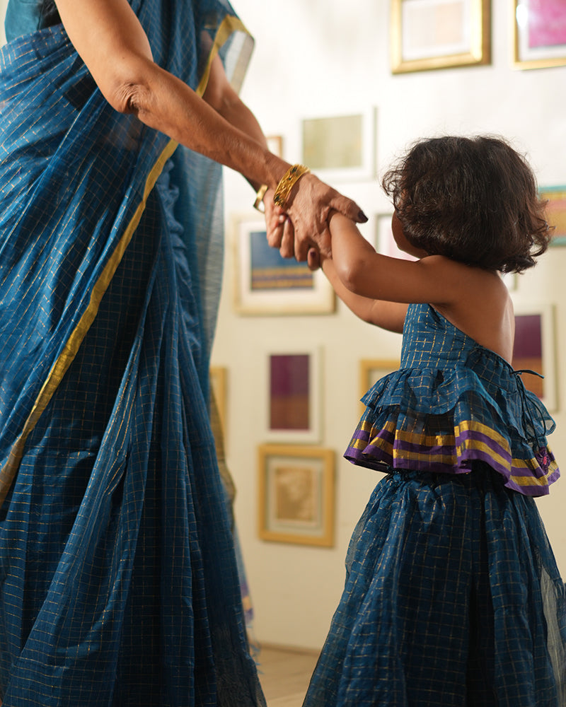 A woman wearing a deep teal blue colored cotton silk saree with golden checks handwoven by weavers in Maheshwar, Madhya Pradesh. This Maheshwari Cotton silk saree is perfect for festivities like Navratri, durga pooja, Dussehra, dushehra, diwali pooja, bhai dooj and weddings. Women's silk wear, silk saree for women, cotton silk saree, heavy pallu border sarees. Nani poti duo. Nani with granddaughter playing. Diwali celebration.