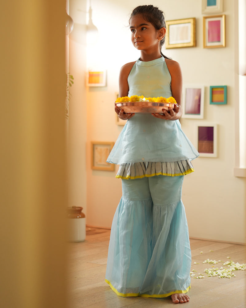 A girl wearing a cute light baby blue and yellow cotton silk sharara suit set handwoven with silver polka dots by weavers in Maheshwar, Madhya Pradesh. This Maheshwari Cotton silk suit set is perfect for festivities like Navratri, durga pooja, Dussehra, dushehra, diwali pooja, bhai dooj and weddings. Kids ethnic outfits, traditional clothing for kids. Silkwear for kids. Halter neck kurti top, salwar kamiz. 