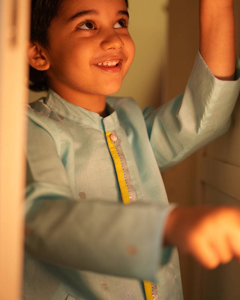 A Boy wearing a light baby blue Indian festive Kurta with golden polka dot motifs made with Handwoven cotton silk fabric woven by artisans in Maheshwar Madhya Pradesh. This kids ethnic kurta is best for upcoming festivities like kids Navratri outfit, kids durga pooja kurta, Dussehra, dushehra, ethnic dress for Diwali, bhai dooj and kids/ boys kurta for weddings. Paired with an off white dhoti/ mundu. Perfect Kids silk wear, ethnic wear for kids, festive clothing for kids.
