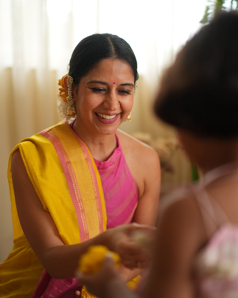 A woman wearing a bright yellow colored cotton silk saree with a pink and gold border and pallu with pink and gold stripes handwoven by weavers in Maheshwar, Madhya Pradesh. This Maheshwari Cotton silk saree is perfect for festivities like Navratri, durga pooja, Dussehra, dushehra, diwali pooja, bhai dooj and weddings. Women's silk wear, silk saree for women, cotton silk saree, heavy pallu sarees. 
