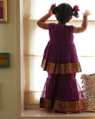 A girl wearing a cute deep purple violet cotton silk sharara suit set handwoven with golden border by weavers in Maheshwar, Madhya Pradesh. This Maheshwari Cotton silk suit set is perfect for festivities like Navratri, durga pooja, Dussehra, dushehra, diwali pooja, bhai dooj and weddings. Kids ethnic outfits, traditional clothing for kids. Silkwear for kids. Sharara set, suit set, salwar set for girls. 