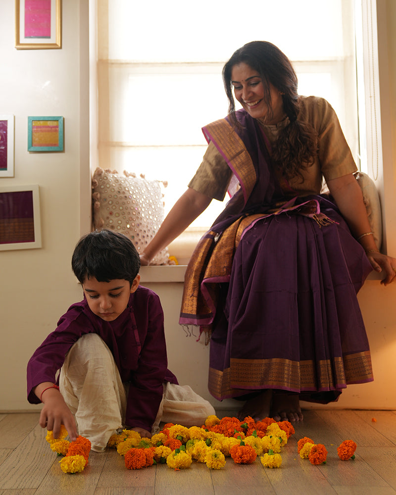 A woman wearing a deep purple wine colored cotton silk saree handwoven by weavers in Maheshwar, Madhya Pradesh. This Maheshwari Cotton silk saree is perfect for festivities like Navratri, durga pooja, Dussehra, dushehra, diwali pooja, bhai dooj and weddings. Mom son duo, mother and son playdate. Mom and son celebrating diwali. 